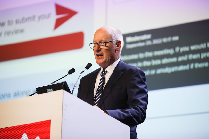 A man with glasses and a navy blue suit and tie stands behind a lecturn at a business conference.